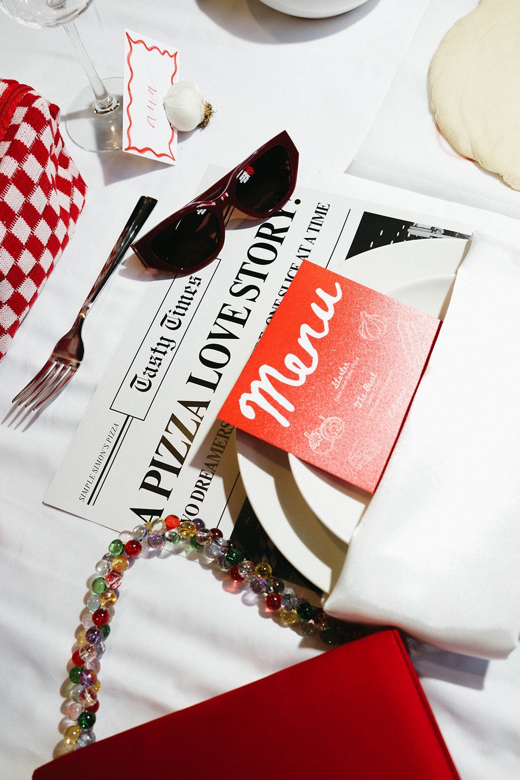 Tasty Times newspaper, red Menu card, sunglasses and a beaded bag styled on the table